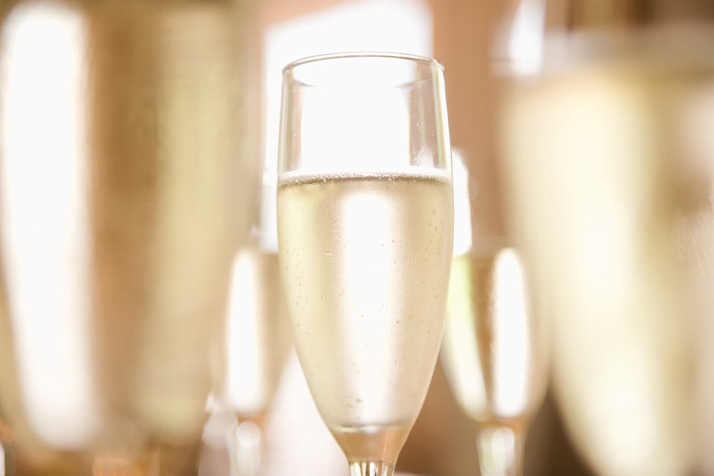 A close-up of a glass of champagne filled with pale sparkling wine, surrounded by blurred glasses in the background. The image conveys a festive or celebratory atmosphere.