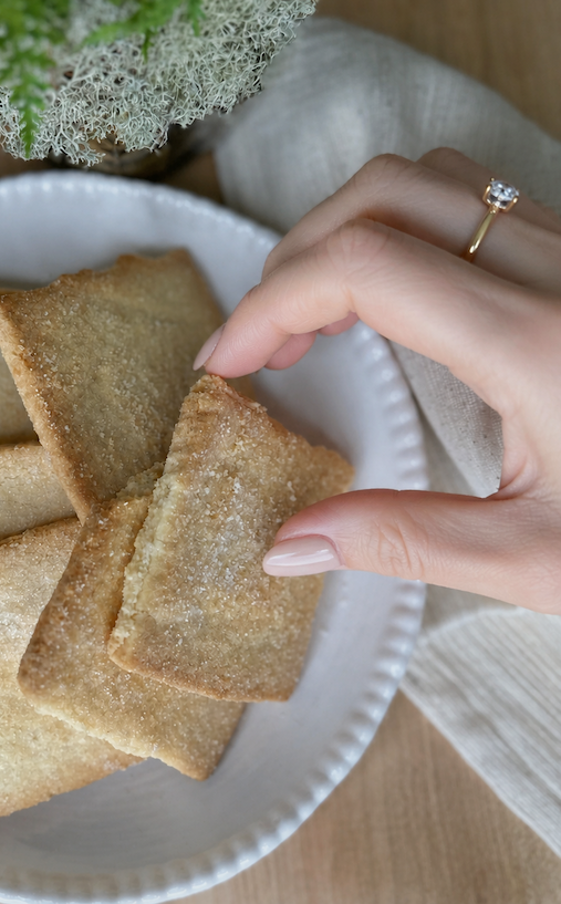 A manicured hand with a delicate gold ring reaches for a sugar-dusted shortbread cookie from a white ceramic plate. Soft neutral linens and natural textures create a warm, elegant kitchen scene.