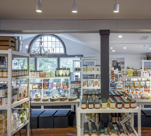 Bright specialty food market with white display shelves stocked with gourmet pantry items, preserves, crackers, sauces, and gifts. Sunlight streams through an arched window at the back, creating an inviting, airy shopping space with warm wood floors and neatly organized merchandise. Fearringotn Village.