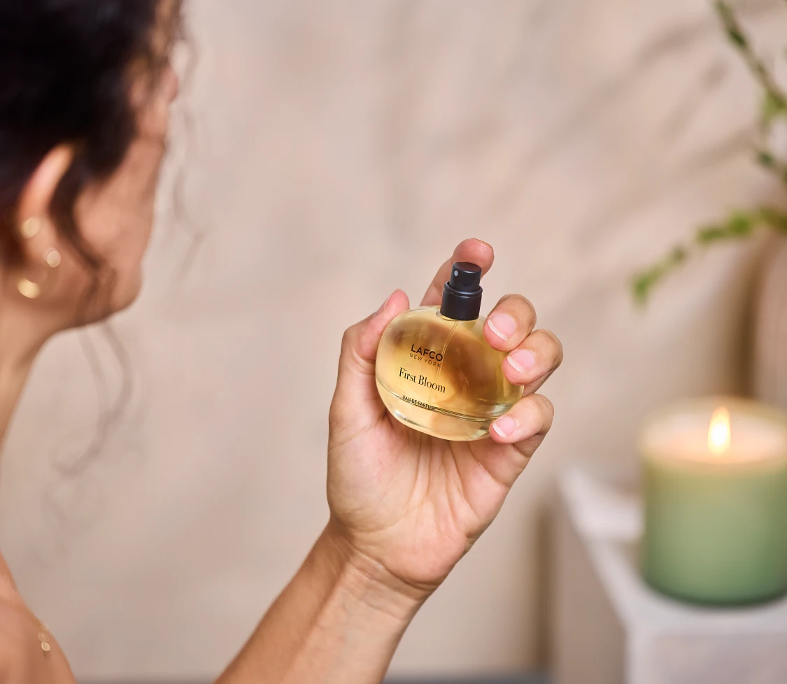Woman holding a bottle of LAFCO First Bloom eau de parfum beside a softly glowing candle. Fearrington Village.