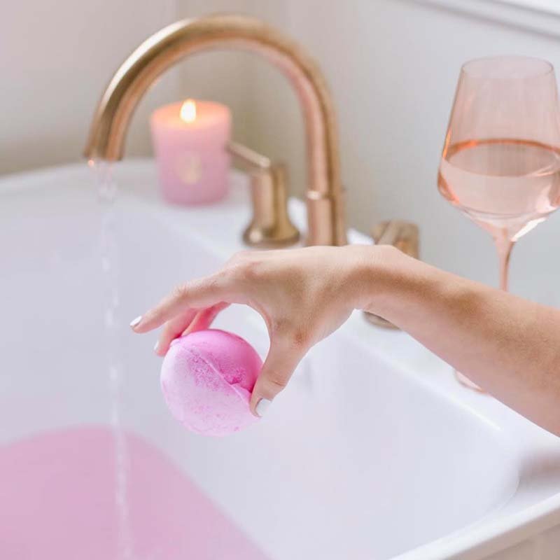 A hand holds a pink bath bomb over a white bathtub, with a glowing candle and a glass of rosé creating a relaxing bath scene. Fearrington Village.