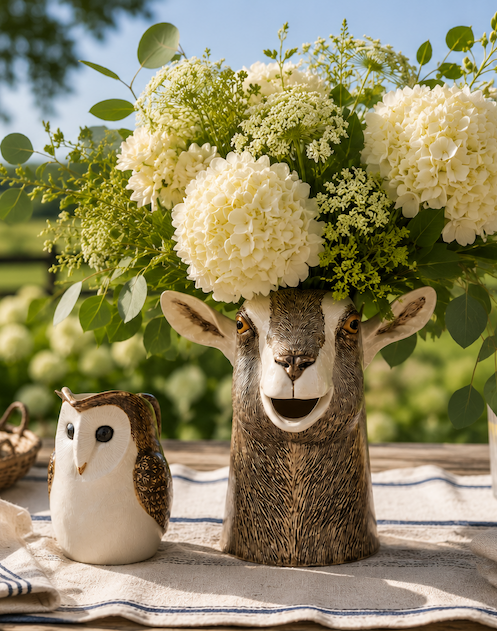 Goat-shaped ceramic vase overflowing with white hydrangeas, Queen Anne's lace, and fresh greenery, paired with a small owl ceramic jug on a linen-covered farmhouse table overlooking a sunlit countryside landscape.