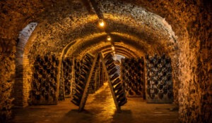 Dimly lit wine cellar with arched stone ceilings, walls, and floors, lined with racks holding many bottles of wine at an angle; yellow lights create a warm, rustic atmosphere. Fearrington Village