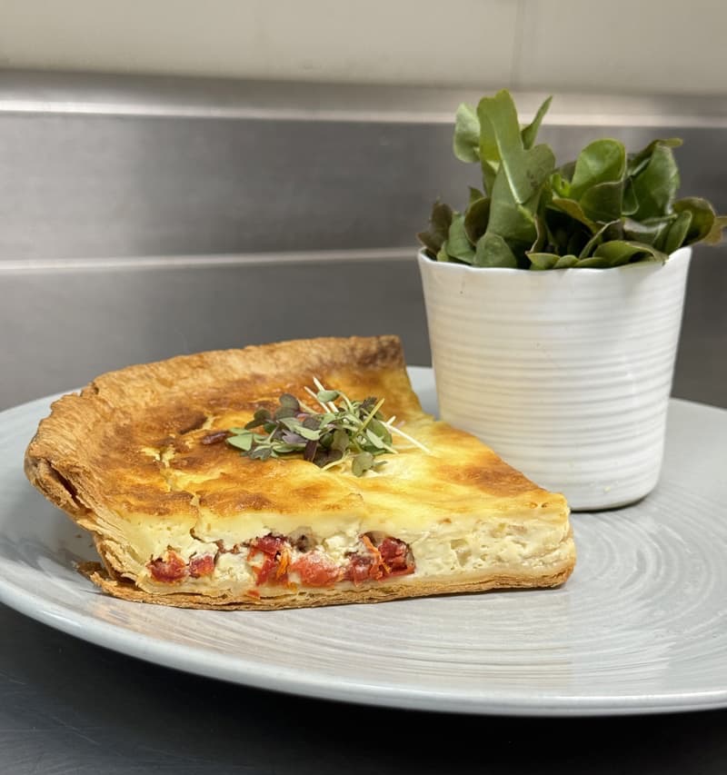 Slice of golden, flaky quiche with visible layers of pastry and a savory filling, topped with fresh microgreens and served on a light gray plate beside a small potted succulent on a stainless steel surface.
