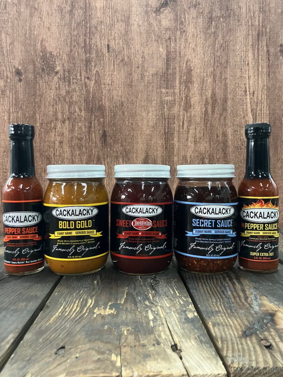 Five Cackalacky sauces and condiments lined up on a rustic wooden table against a weathered wood backdrop. The assortment includes Original Pepper Sauce, Bold Gold mustard, Sweet Cheerwine Sauce, Secret Sauce, and Super Extra Hot Pepper Sauce, showcasing a range of flavors from tangy and sweet to intensely spicy.