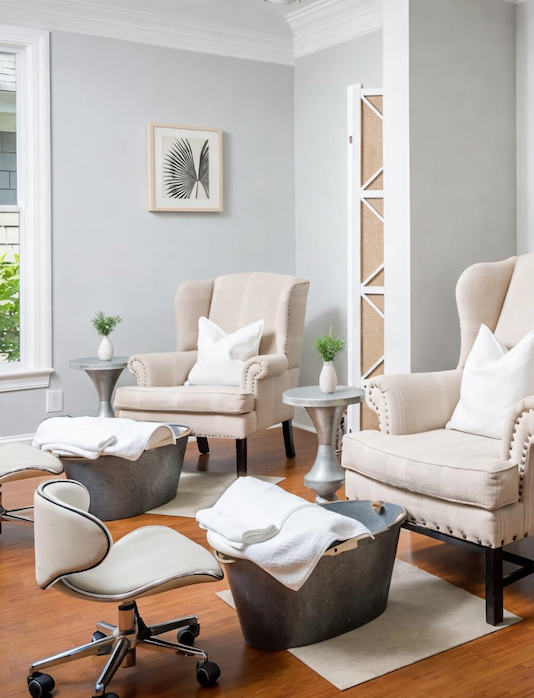Bright, serene spa pedicure room with two upholstered armchairs, foot basins with neatly folded white towels, rolling stools, and soft neutral décor accented by framed botanical prints and warm wood floors.