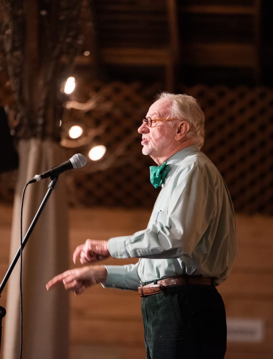 An older man in glasses and a green bow tie speaks into a microphone onstage, gesturing with his hands under warm lighting.