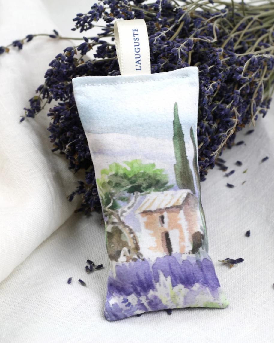 A small fabric lavender sachet rests against a bundle of dried lavender stems, with a soft watercolor design of a rustic house, trees, and purple fields. A simple fabric tag at the top reads “L’AUGUSTE,” and a few loose lavender buds are scattered nearby on a light linen surface.