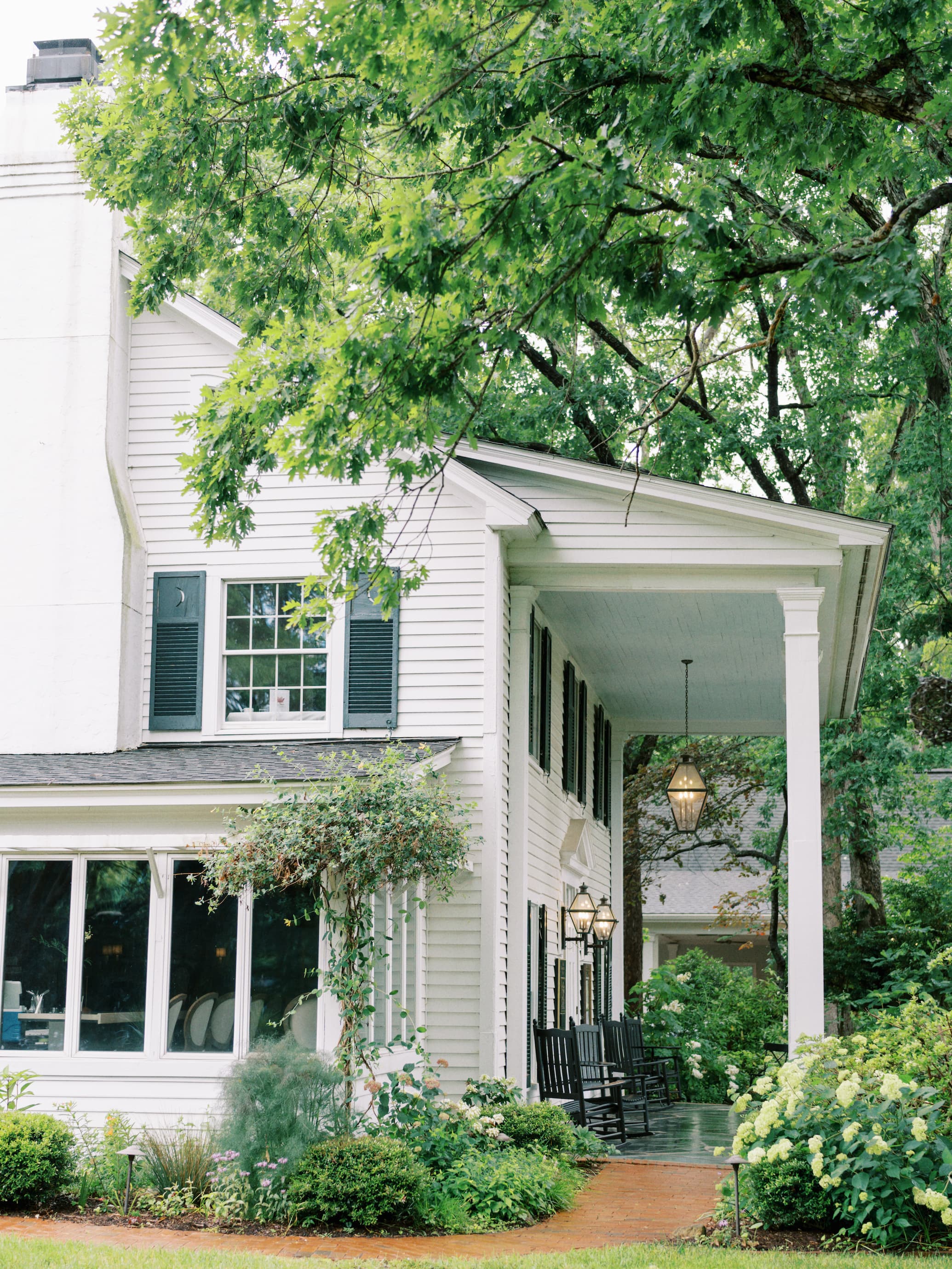 Classic white clapboard inn surrounded by lush summer gardens, featuring a wide covered porch with rocking chairs, brick walkways, blooming hydrangeas, and mature shade trees.