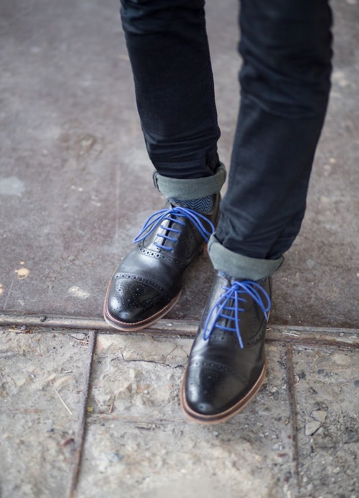 A person wearing black leather brogue shoes with bright blue laces, standing on a stone and concrete surface with jeans cuffed at the ankles.