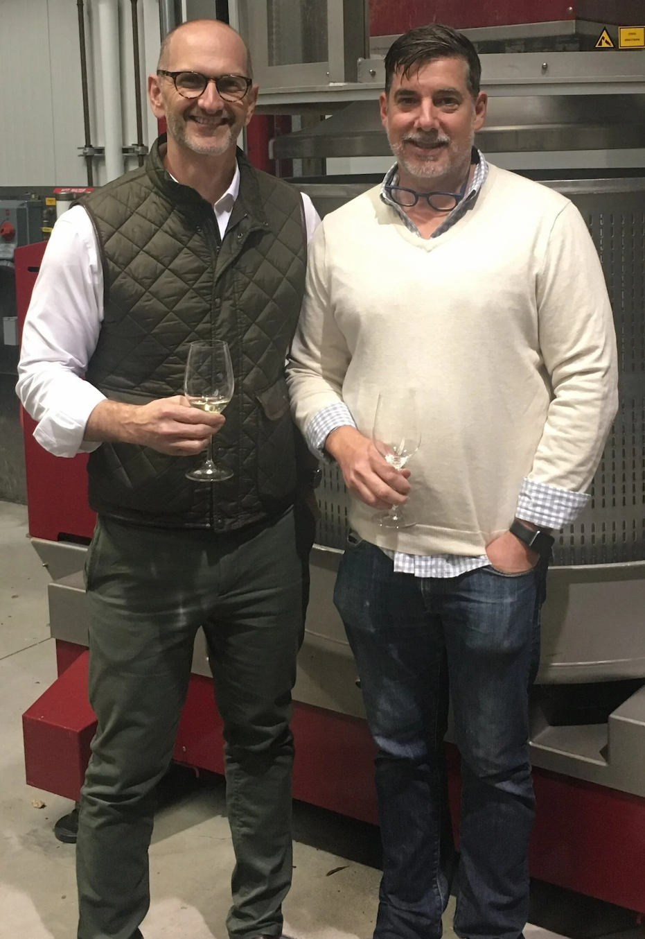 Two men holding wine glasses stand together in a winery production space, dressed in smart casual attire with stainless steel winemaking equipment in the background. Fearrington Village.