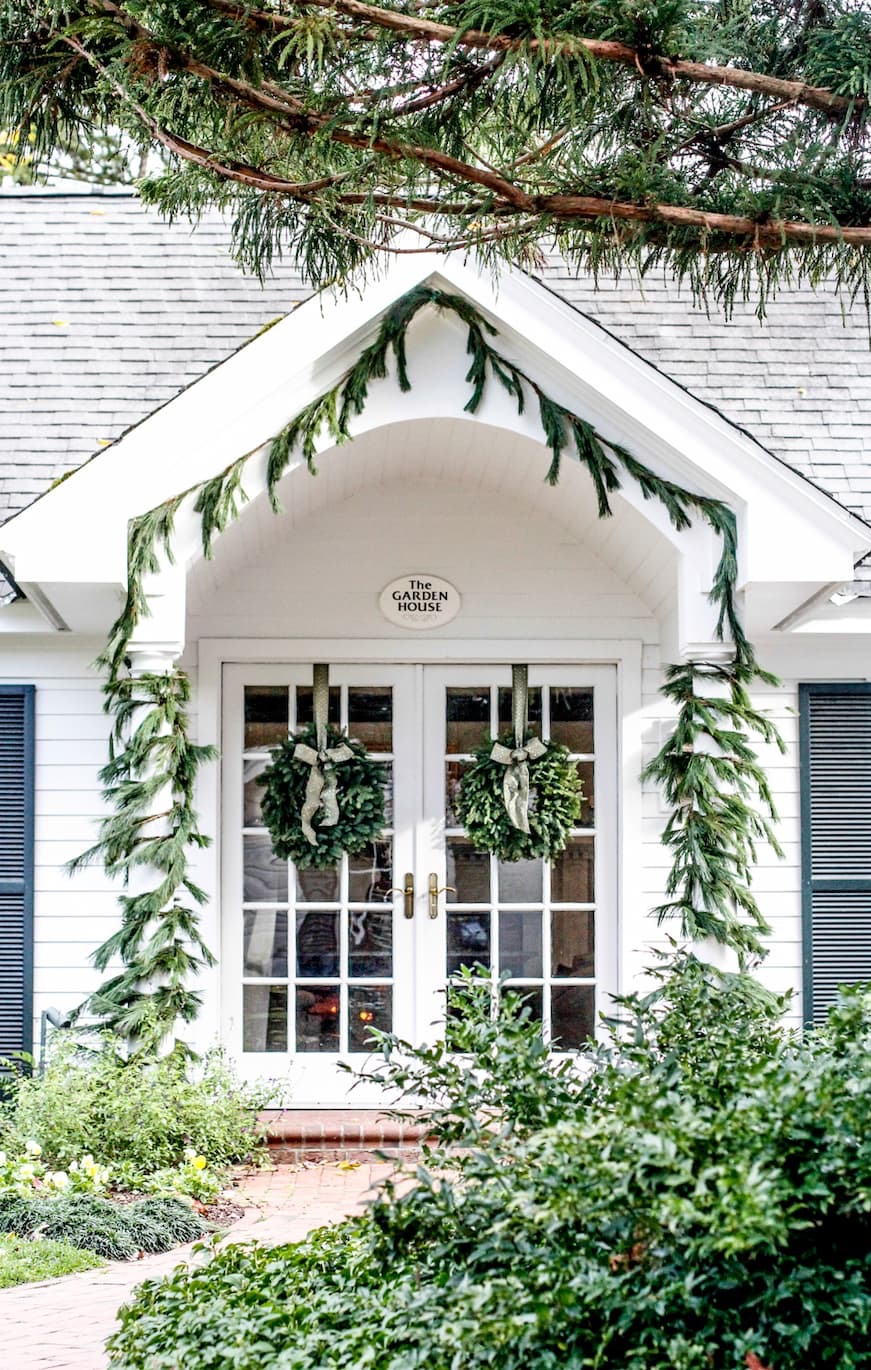 A white house entrance with double French doors decorated with two wreaths and draped evergreen garlands, surrounded by greenery and brick steps leading to the door. Fearrington Village
