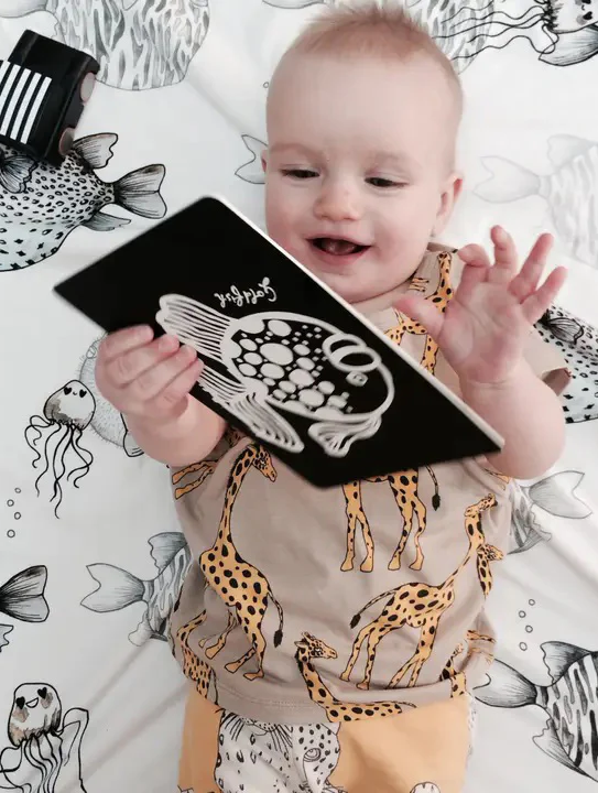 Smiling baby lying on a patterned blanket, holding a small black board book with a white animal illustration, wearing a tan outfit printed with giraffes.