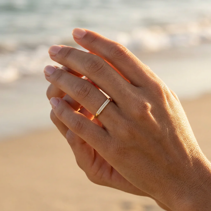 Close-up of a hand wearing a slim gold pavé diamond band, photographed in warm natural light with a softly blurred beach and ocean backdrop, highlighting the ring’s understated sparkle and elegant design.