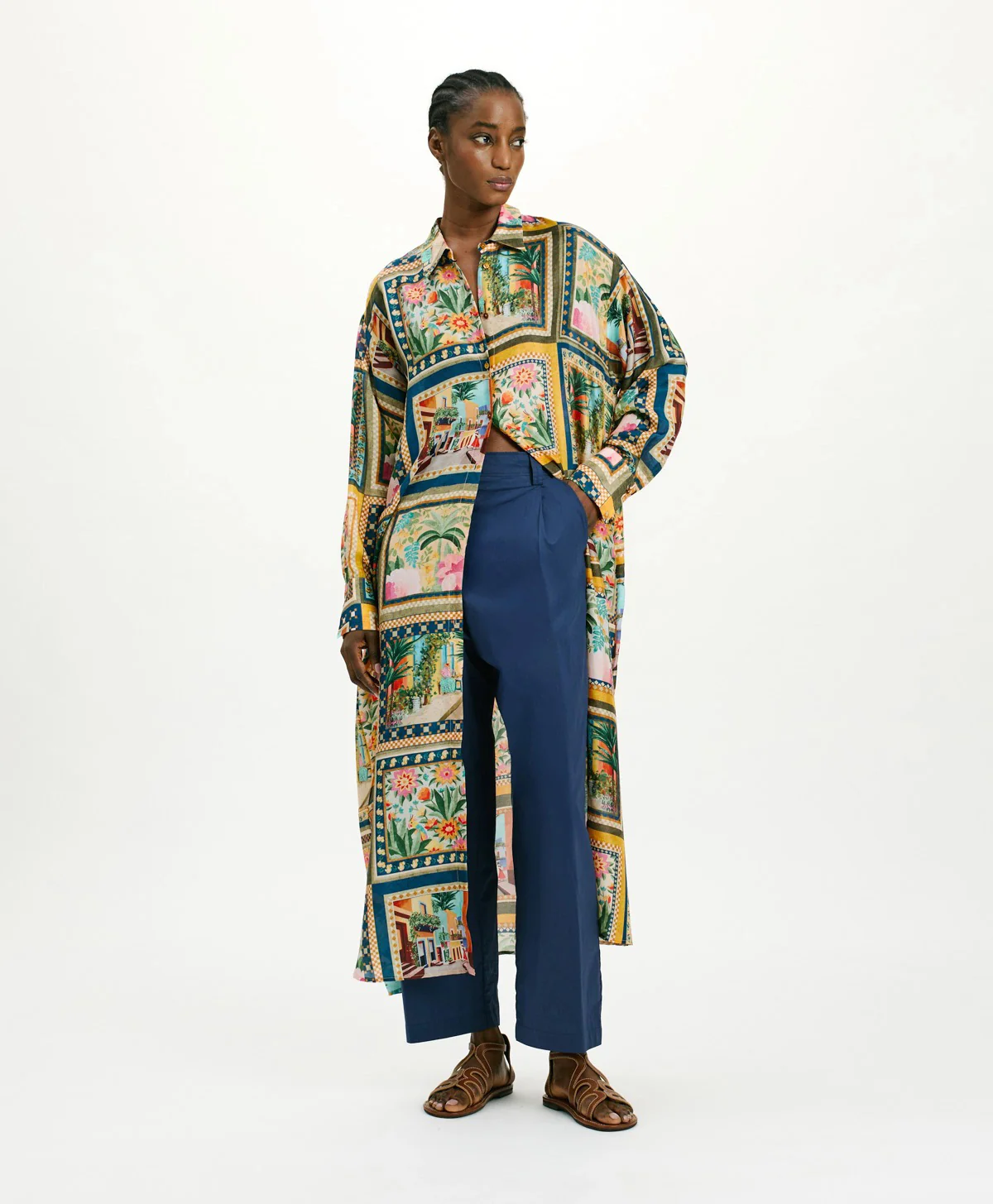A woman stands against a white background wearing a long, patchwork-print shirt dress layered over navy wide-leg pants. The dress features colorful scenic and floral panels, and she pairs the look with brown leather sandals, one hand in her pocket as she looks to the side.