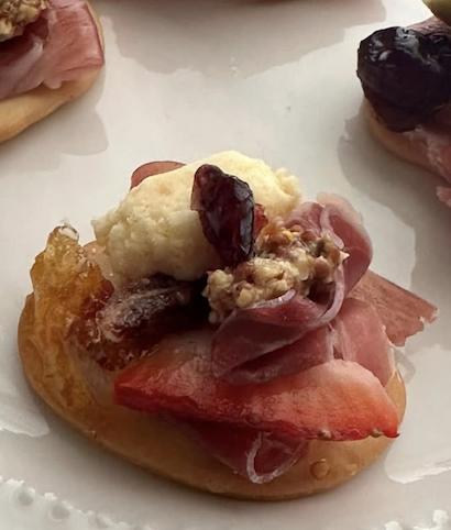 A close-up of an appetizer on a white plate, featuring a small toasted round topped with thinly sliced prosciutto, a dollop of creamy cheese, a date, and a sprinkle of nuts. Other similar bites are softly visible in the background.