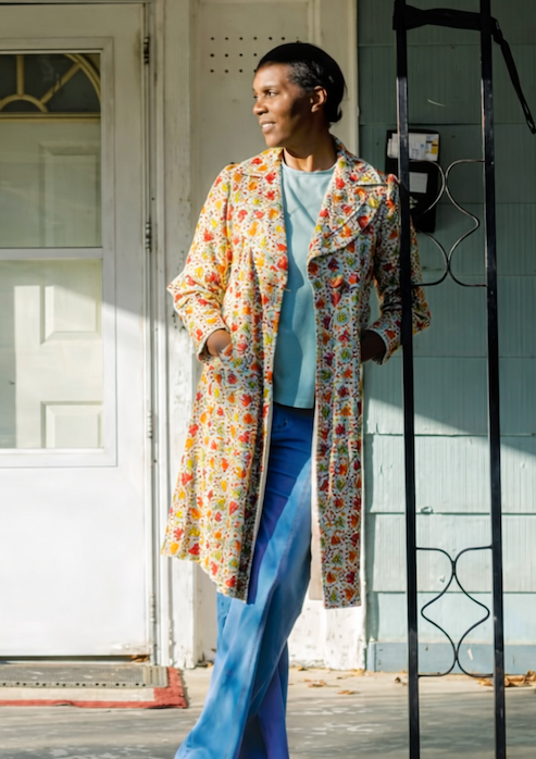 Spring Council wearing a long floral coat and blue trousers, standing on a sunlit porch in front of a pale green house with a white door.