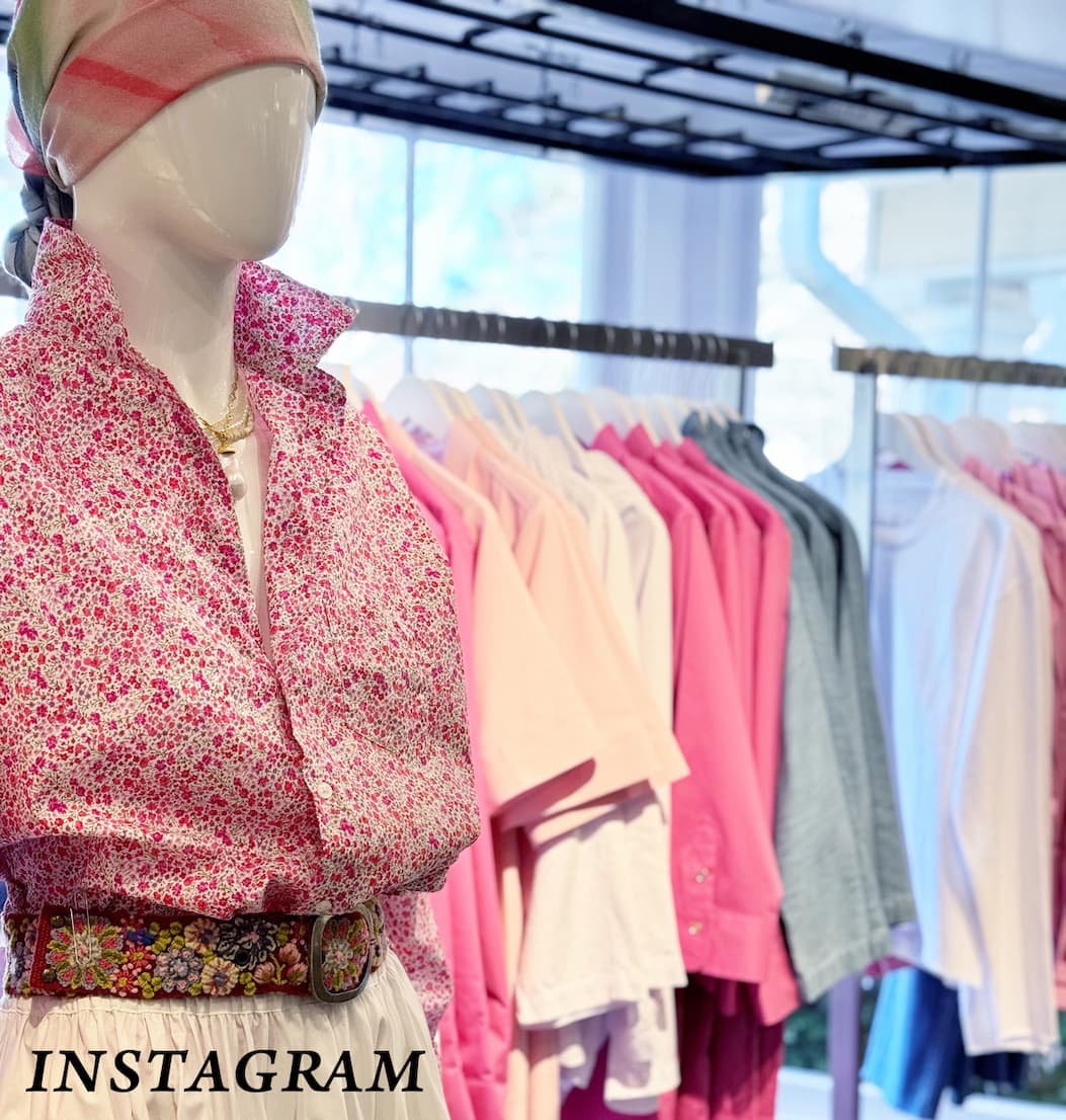 Dovecote mannequin dressed in a pink floral blouse with a patterned belt and light skirt, styled with a soft headscarf, standing in front of a clothing rack filled with pink, white, and pastel garments inside a boutique.