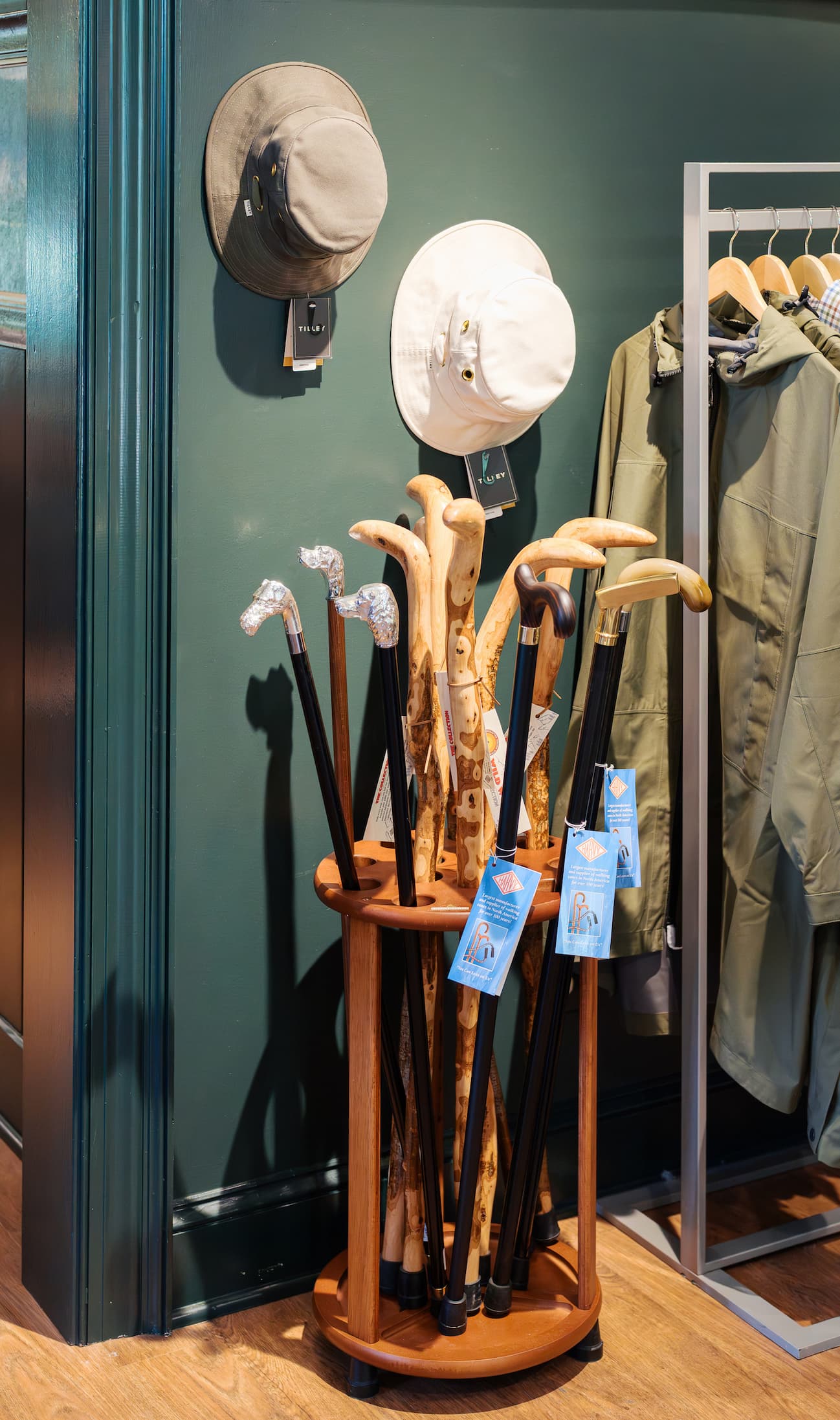 A display stand holds a variety of walking canes with different handle styles, set against a dark green wall. Above them, two hats hang on hooks, and a clothing rack with jackets stands nearby, creating a refined retail setting.