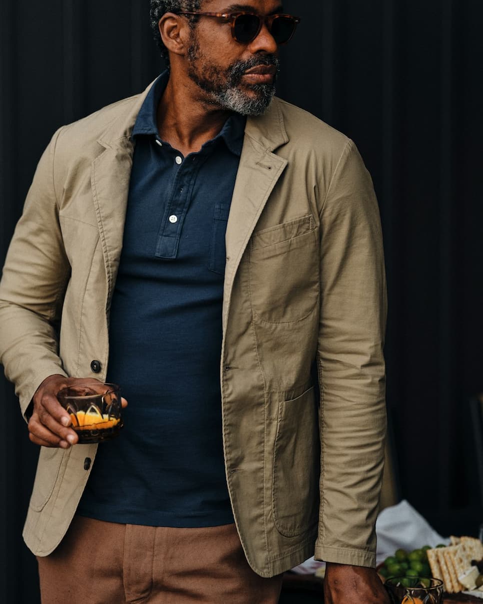 A man wearing sunglasses, a tan casual blazer and navy polo holds a cocktail against a dark backdrop. Fearrington Village.