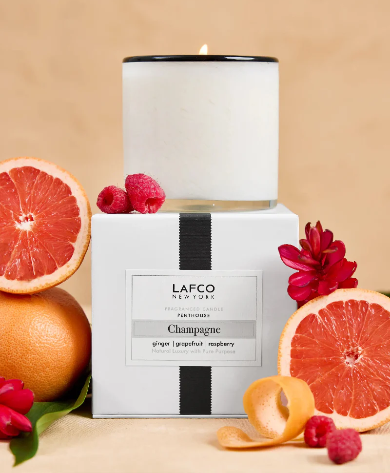 White glass candle labeled “LAFCO New York Champagne,” displayed on its box with grapefruit halves, raspberries, and red flowers against a warm background.