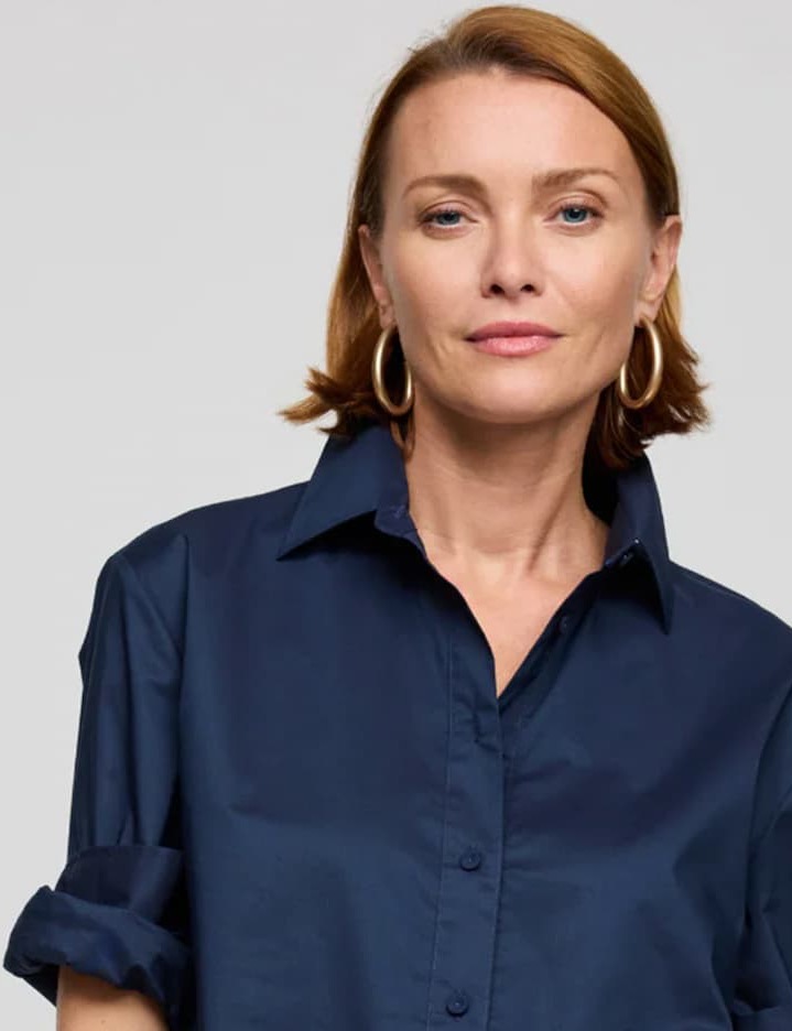 Woman with short auburn hair wearing a navy button-down shirt and gold hoop earrings against a light background.