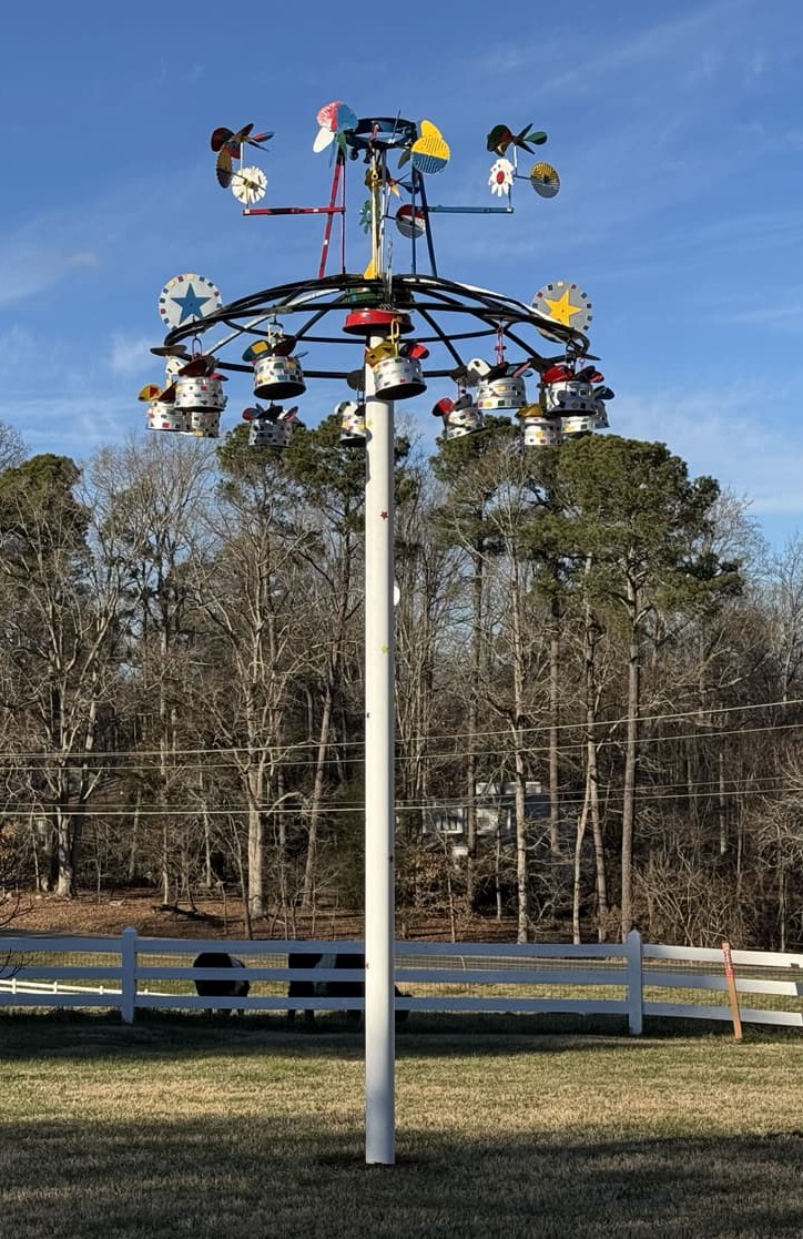 Tall outdoor kinetic sculpture with a white pole and a circular, colorful metal carousel at the top, featuring small spinning shapes, bells, and star motifs against a blue sky, with a fenced pasture and trees in the background.