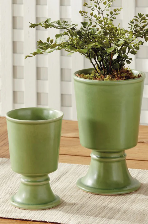 Two green glazed footed cachepots on a table, one holding a small leafy plant with moss, styled indoors against a light paneled backdrop.
