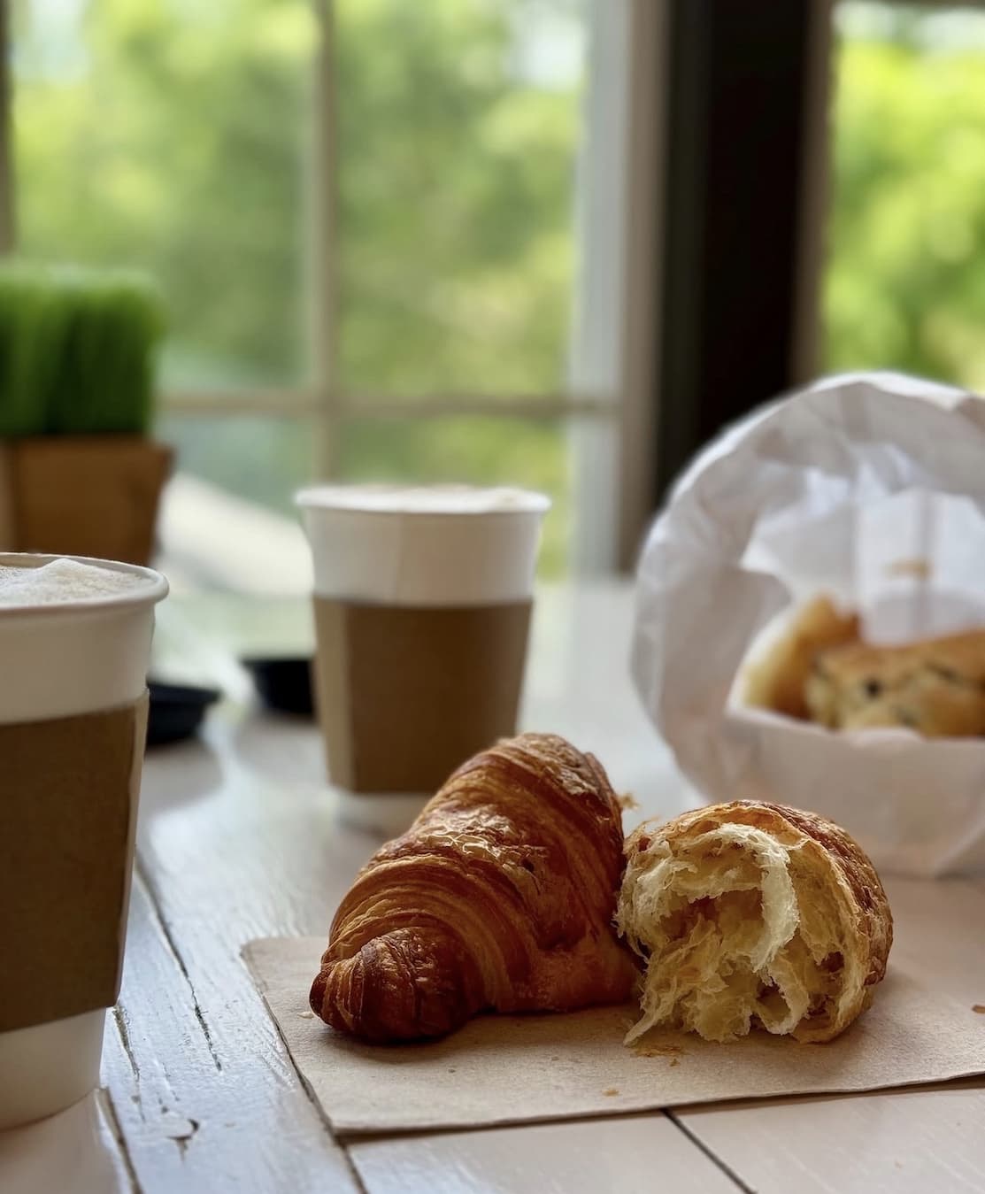 Fresh croissants and frothy coffee sit on a rustic wooden table beside a sunlit window. The warm pastries, soft natural light, and casual café setting create a cozy morning breakfast scene.