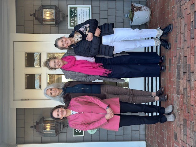 Four women stand on a brick entryway in front of a shingled building entrance, flanked by lantern lights and a sign reading “Fitch Creations, Inc.,” with one woman holding a small dog.