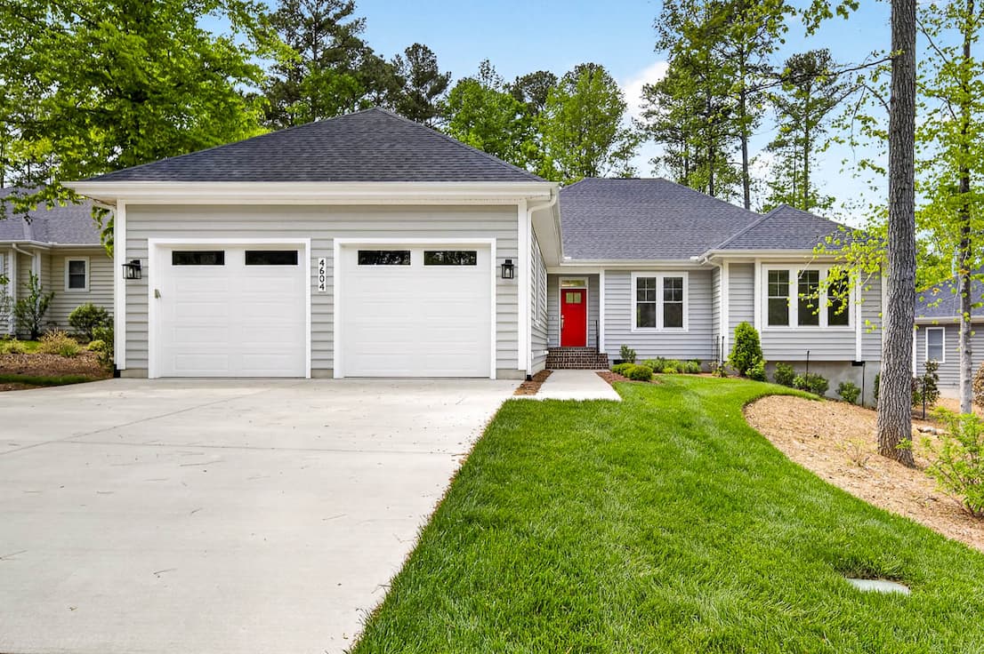 A single-story home with gray siding and a red front door features a two-car garage, a wide driveway, and a neatly maintained lawn, set among tall trees in a quiet neighborhood.