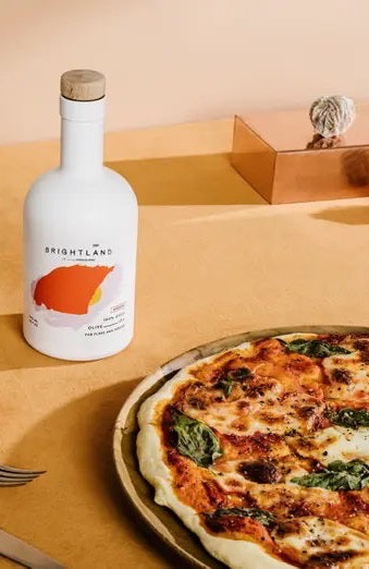 A Margherita-style pizza with melted cheese and basil sits beside a bottle of Brightland olive oil on a warm-toned surface, styled with clean, modern accents.