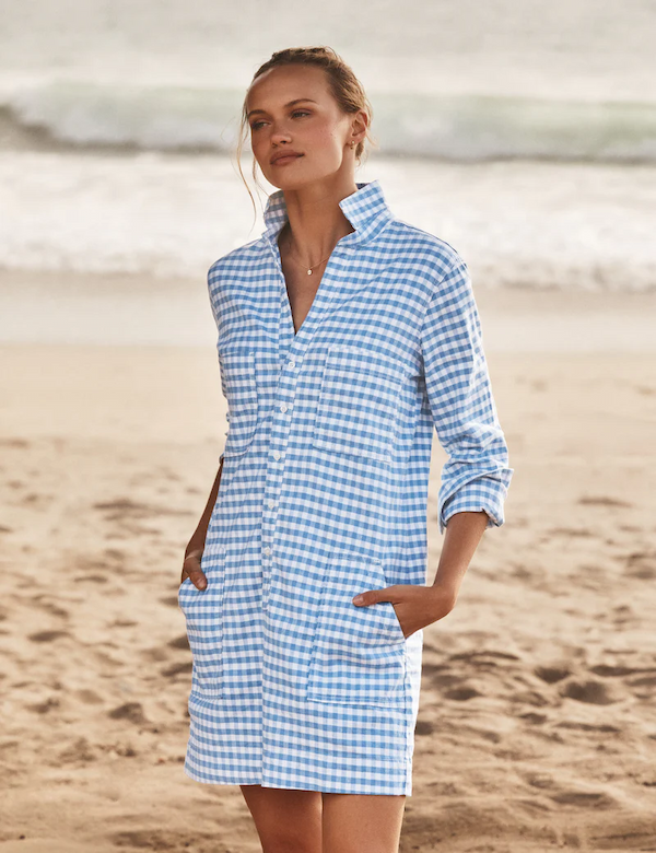 Woman standing on a sandy beach wearing a relaxed, blue-and-white gingham shirt dress with a popped collar and front pockets. Hands in pockets, with soft waves and shoreline in the background, creating a breezy, coastal feel.