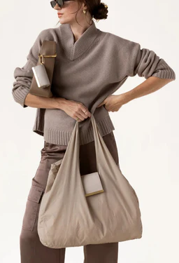 A stylish woman carries an oversized taupe tote bag over one arm while dressed in a monochromatic neutral outfit. She wears a soft draped knit sweater, coordinating trousers, and oversized sunglasses, creating an effortlessly modern look. Attached to the tote and a structured shoulder bag are matching leather accessories with gold-tone hardware, highlighting the collection's blend of practicality and refined design.