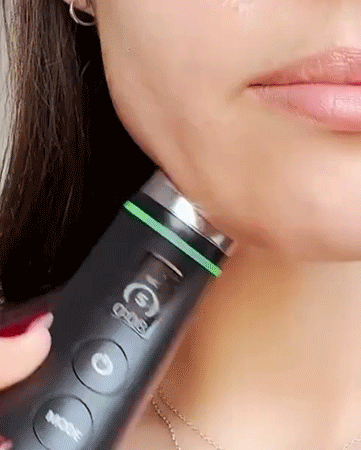 Close-up of a woman’s jawline as she uses a handheld skincare device with a metal tip and illuminated ring along her chin.