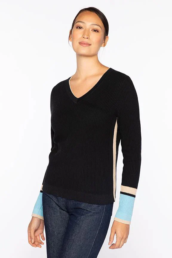 Woman wearing a black ribbed V-neck sweater with light blue and cream color-blocked cuffs, paired with dark jeans, standing against a white background.