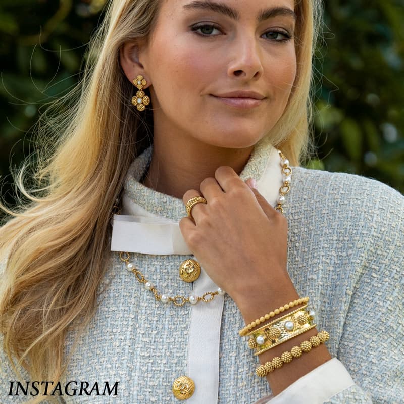 Blonde woman in a textured tweed jacket wearing gold and pearl earrings, layered necklaces, rings, and stacked bracelets, posed outdoors.