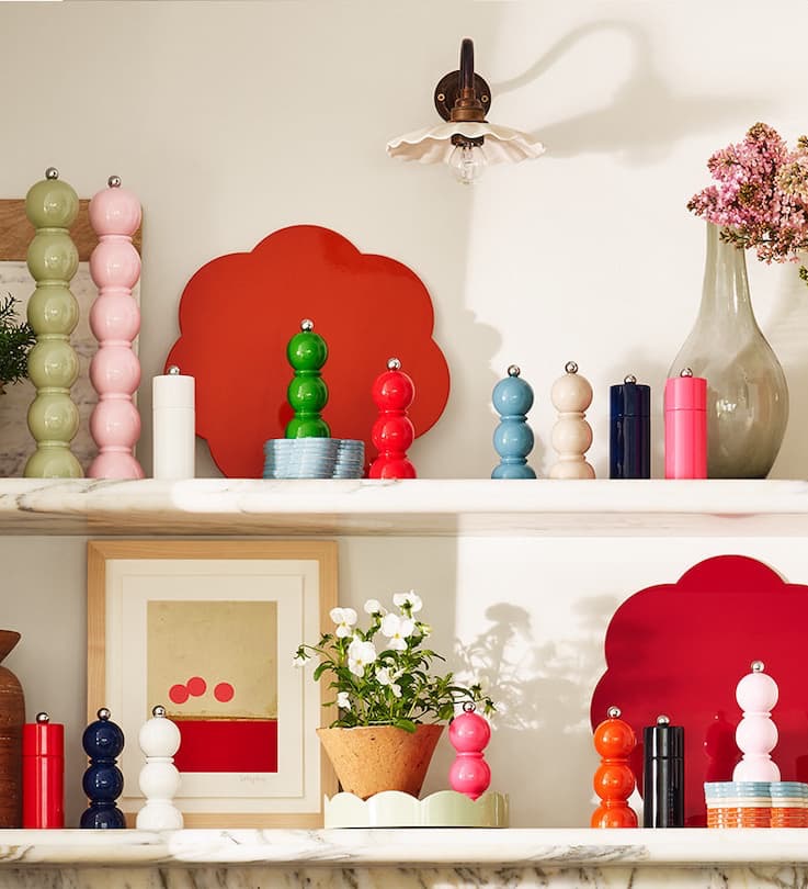 Playful, color-saturated shelf display featuring glossy pepper mills in pastel and bold hues, layered with sculptural red backplates, ceramics, framed art, and fresh flowers; styled on marble shelving with a mix of symmetry and whimsy.
