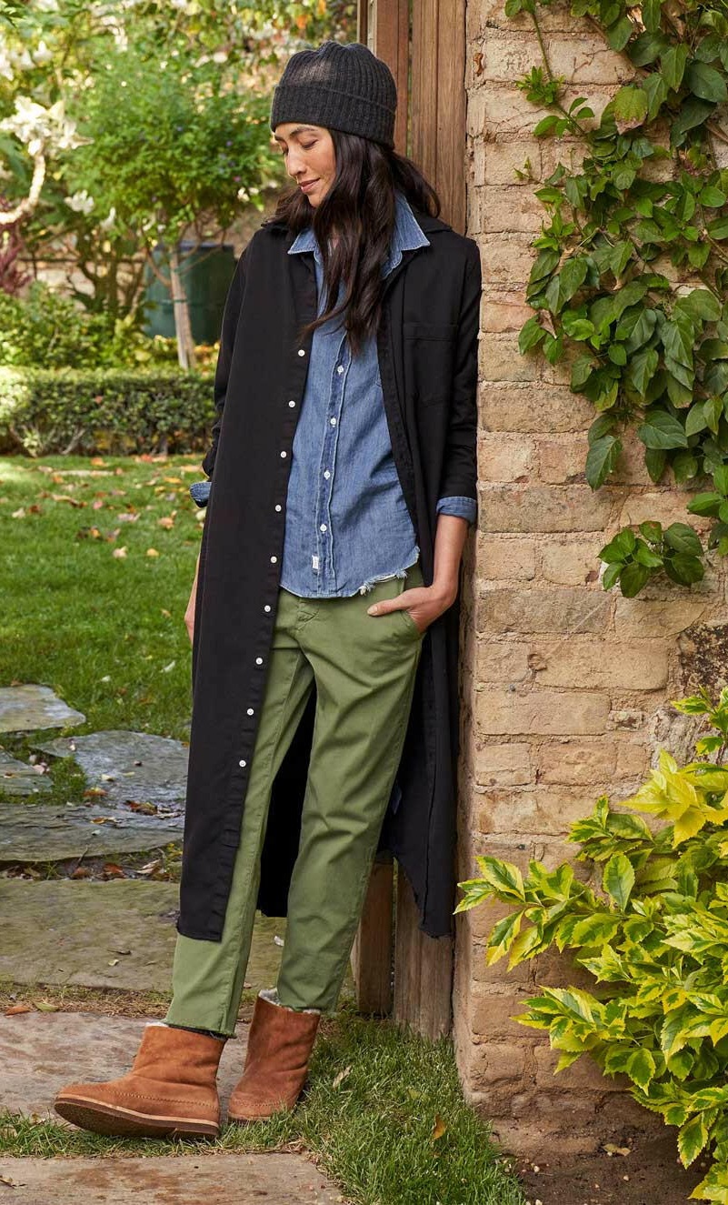 A woman wearing a black long coat, blue denim shirt, green pants, and brown boots leans against a brick wall outdoors.