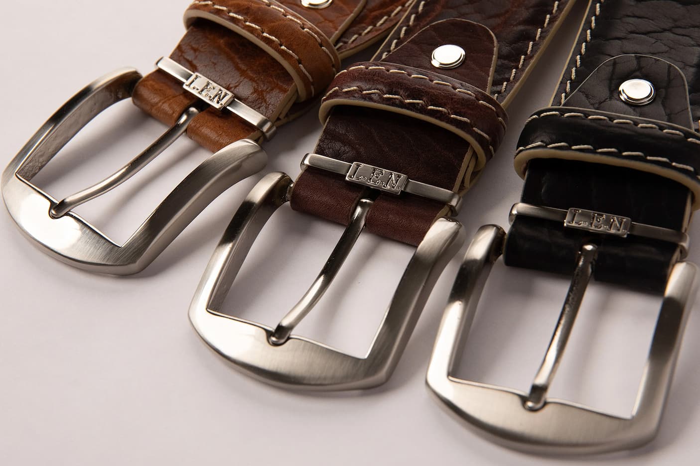 Three leather belts in tan, brown, and black with stitched edges and brushed metal buckles arranged side by side on a neutral background.