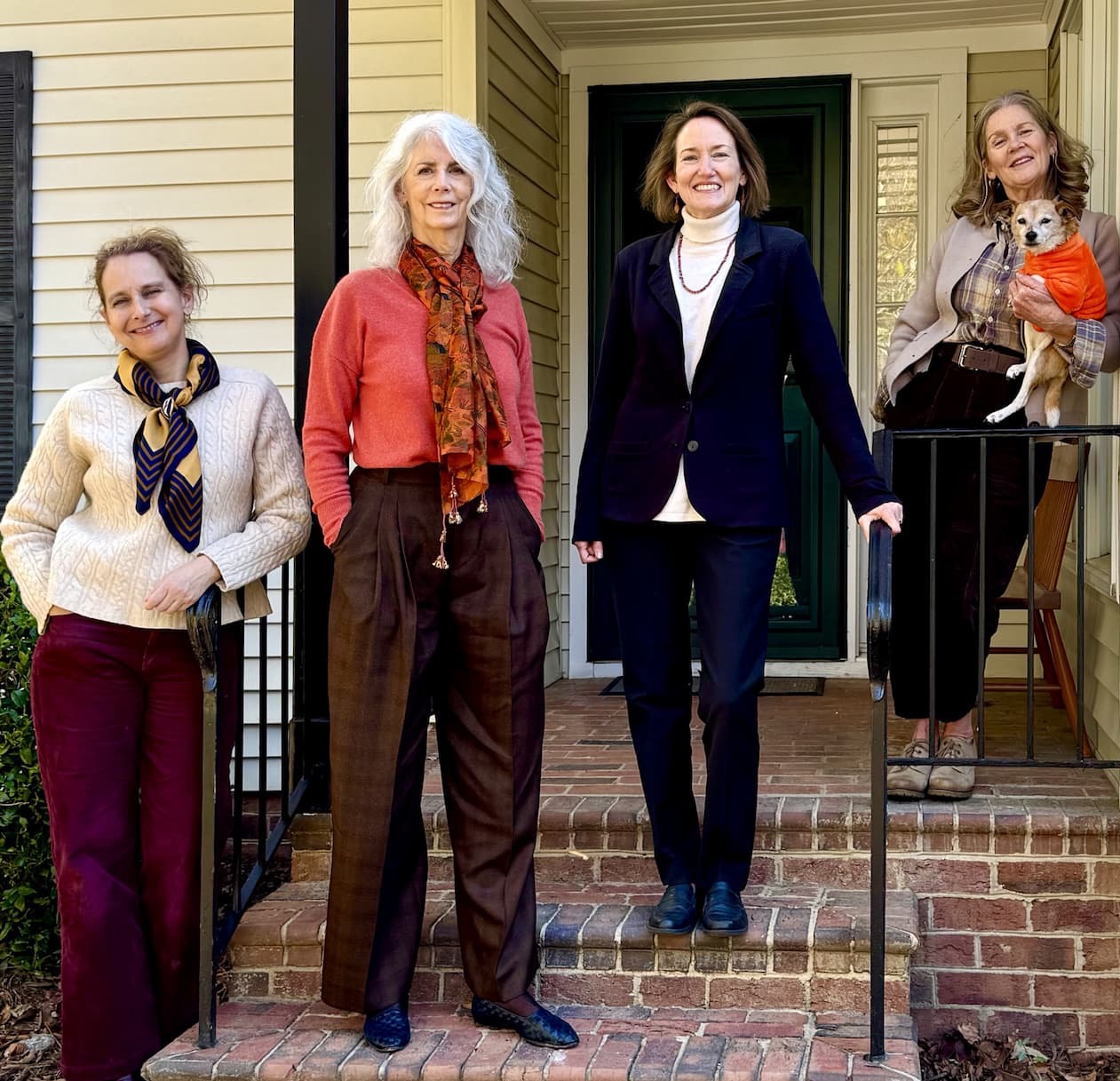 Four women pose together outside a house; three stand on the steps, one holds a small dog wearing an orange sweater. They are smiling and dressed in casual to business-casual attire. Fearrington Village