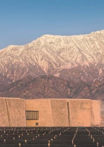 Modern concrete building in the foreground with rows of solar panels, set against a backdrop of snow-dusted mountains at sunset.