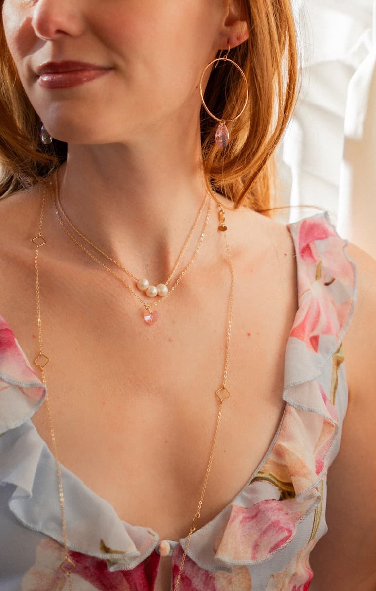 A close-up of a person wearing layered gold necklaces with pearls and a pink pendant, paired with large hoop earrings and a soft floral dress in blush tones.