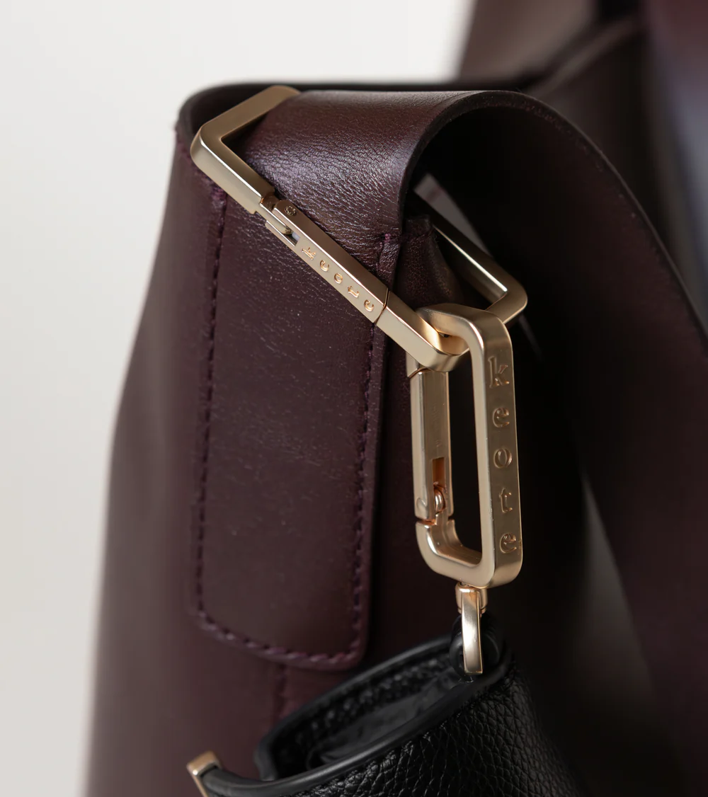 Close-up detail of a dark brown leather handbag featuring polished gold-tone hardware and clasp accents. The image highlights the bag’s smooth leather texture, precise stitching, and elegant metal fastenings against a soft neutral background.