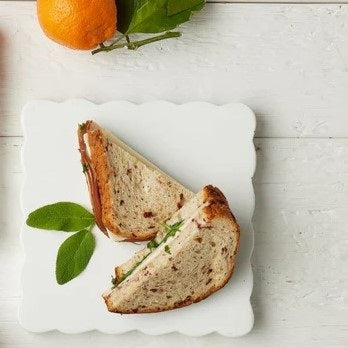 White scalloped serving board styled with two halves of a gourmet sandwich on rustic bread, accented with fresh green leaves and a citrus fruit on a whitewashed tabletop. The clean, minimalist setting highlights the board’s soft curves and elegant presentation.