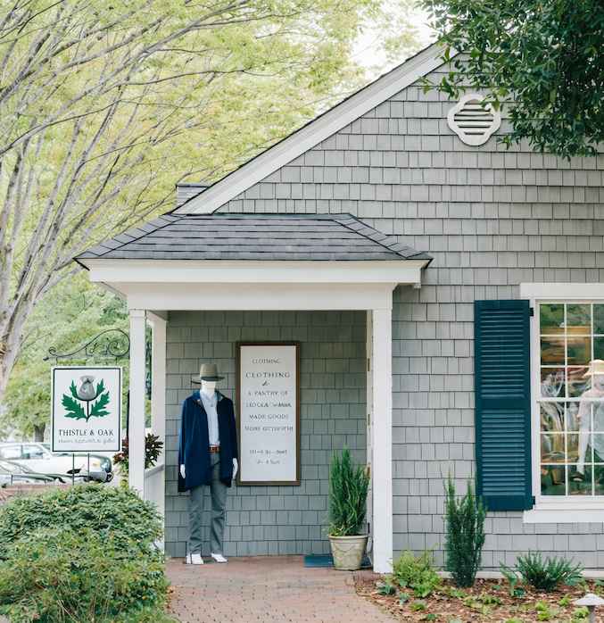 Exterior of a quaint shingled boutique with a small covered porch and green shutters, a Thistle & Oak sign by the walkway, and a mannequin dressed in a jacket and hat standing near the entrance under leafy trees.