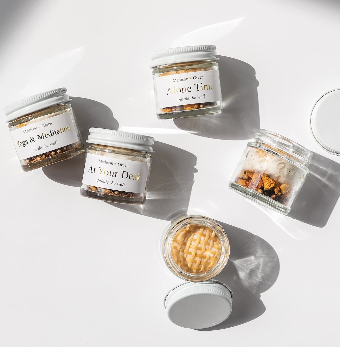 Flat-lay of small glass jars labeled “Alone Time,” “Yoga & Meditation,” and “At Your Desk,” arranged in bright sunlight on a clean white surface, with lids off to reveal textured, botanical contents casting soft shadows.