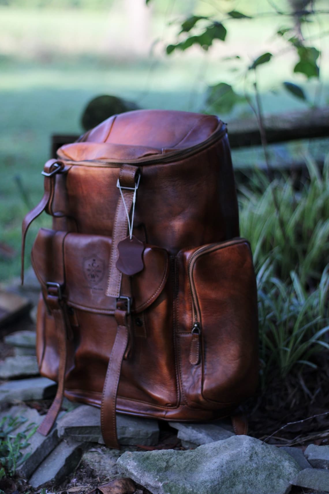 Brown leather backpack with multiple pockets and straps, set on stones in a garden with soft greenery in the background.
