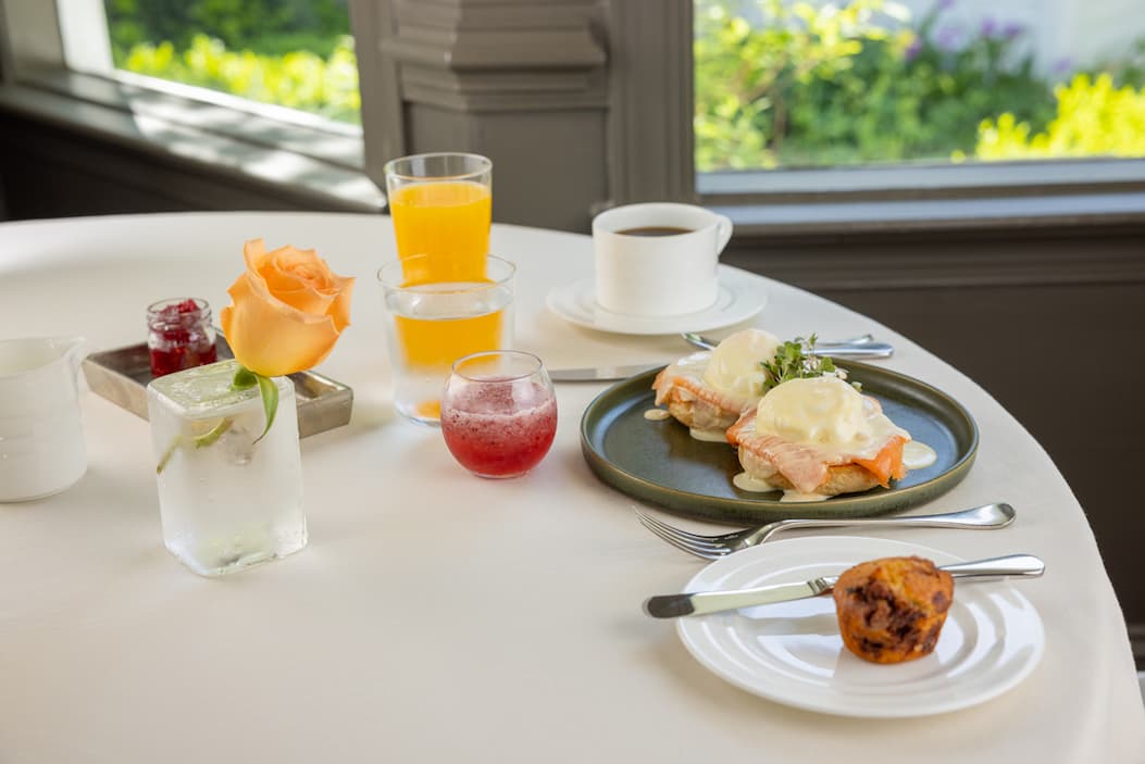 A bright breakfast table is set near large windows overlooking a garden. The meal includes eggs Benedict topped with smoked salmon and hollandaise sauce, a freshly baked muffin, coffee, orange juice, and small glasses of water and berry juice. A single peach-colored rose in a clear glass vase serves as the centerpiece, adding an elegant touch to the relaxed, sunlit dining experience.