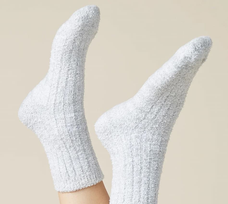 A close-up of two feet wearing soft, fuzzy white crew socks against a light beige background. The plush socks have a cozy ribbed texture and are shown with the toes pointed upward.